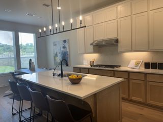 Model Home A modern kitchen with a bright, open layout, sleek black hardware, warm cabinetry, and elegant pendant lighting over a large island.
