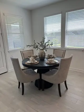 Model Home A cozy dining nook with a round black table, beige upholstered chairs, and bright window blinds.