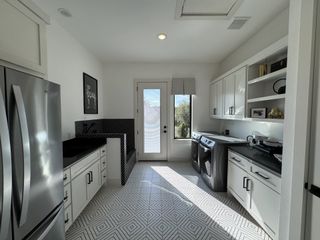 A well-lit laundry room with patterned tile flooring, white cabinetry, a stainless steel refrigerator, and a door leading to the outside.