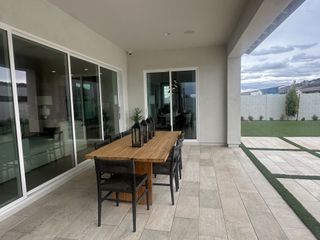 A covered patio features a wooden dining table with black woven chairs, stone tile flooring, and large sliding glass doors leading to an indoor living space, overlooking a landscaped backyard.