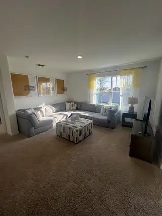 A cozy living area with a large sectional sofa, warm carpeted flooring, and natural light streaming through large windows.