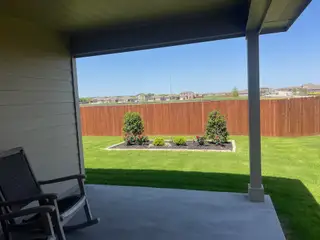 Model Home A cozy patio overlooks a lush green lawn and landscaped garden beds, framed by a wooden fence.
