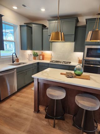 A modern kitchen with teal cabinets, brass pendant lights, a white island, and wood flooring.