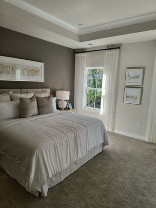 Model Home A cozy bedroom with a plush bed, neutral tones, elegant artwork, and ample natural light through sheer curtains.
