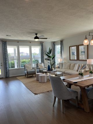 A cozy living area with natural light, a beige sectional, patterned chairs, and a wooden dining table on wood flooring.