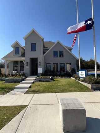 A modern gray home with a manicured lawn and flags in Cross Creek Meadows 55' & 60' Series by Normandy Homes (Celina, TX).
