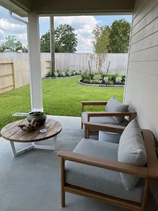 A cozy patio with wooden chairs, gray cushions, and a round table overlooks a lush, fenced backyard with landscaped gardens.