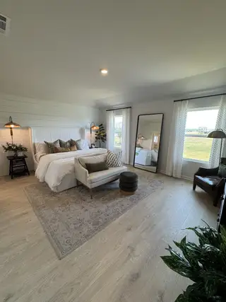 A spacious bedroom with cozy seating, hardwood floors, and soft lighting, featuring elegant décor and large windows.