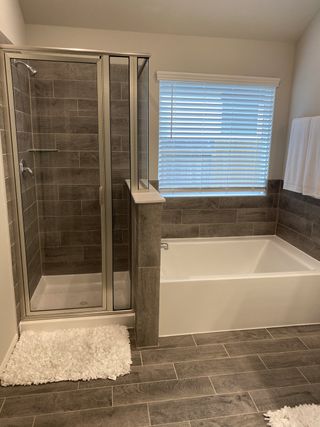 Model Home A modern bathroom with a sleek glass shower, deep bath, textured tiles, and soft natural light through a window.