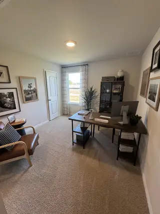 A cozy home office with beige carpet, a wooden desk, and art-adorned walls, offering a serene workspace ambience.