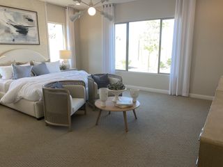 A bright bedroom with neutral carpet features a sitting area with rattan chairs, a plush bed, and large windows, creating a serene retreat.