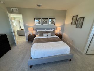 Model Home A well-lit bedroom featuring a contemporary gray upholstered bed with neutral bedding. The walls have framed botanical prints, and two matching nightstands with lamps flank the bed.