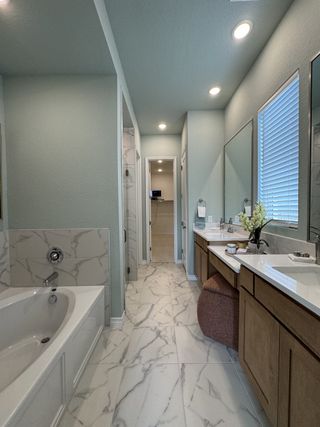 A spacious bathroom with dual vanities, warm wood cabinets, marble-patterned tile floors, and soft blue walls creates an elegant, relaxing space.